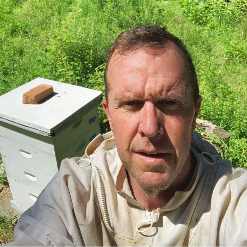 The image shows a man in a beekeeping suit, taking a selfie. He is standing in front of a beehive. The background is filled with green plants. There is a brick on top of the beehive. The man has a serious expression on his face.